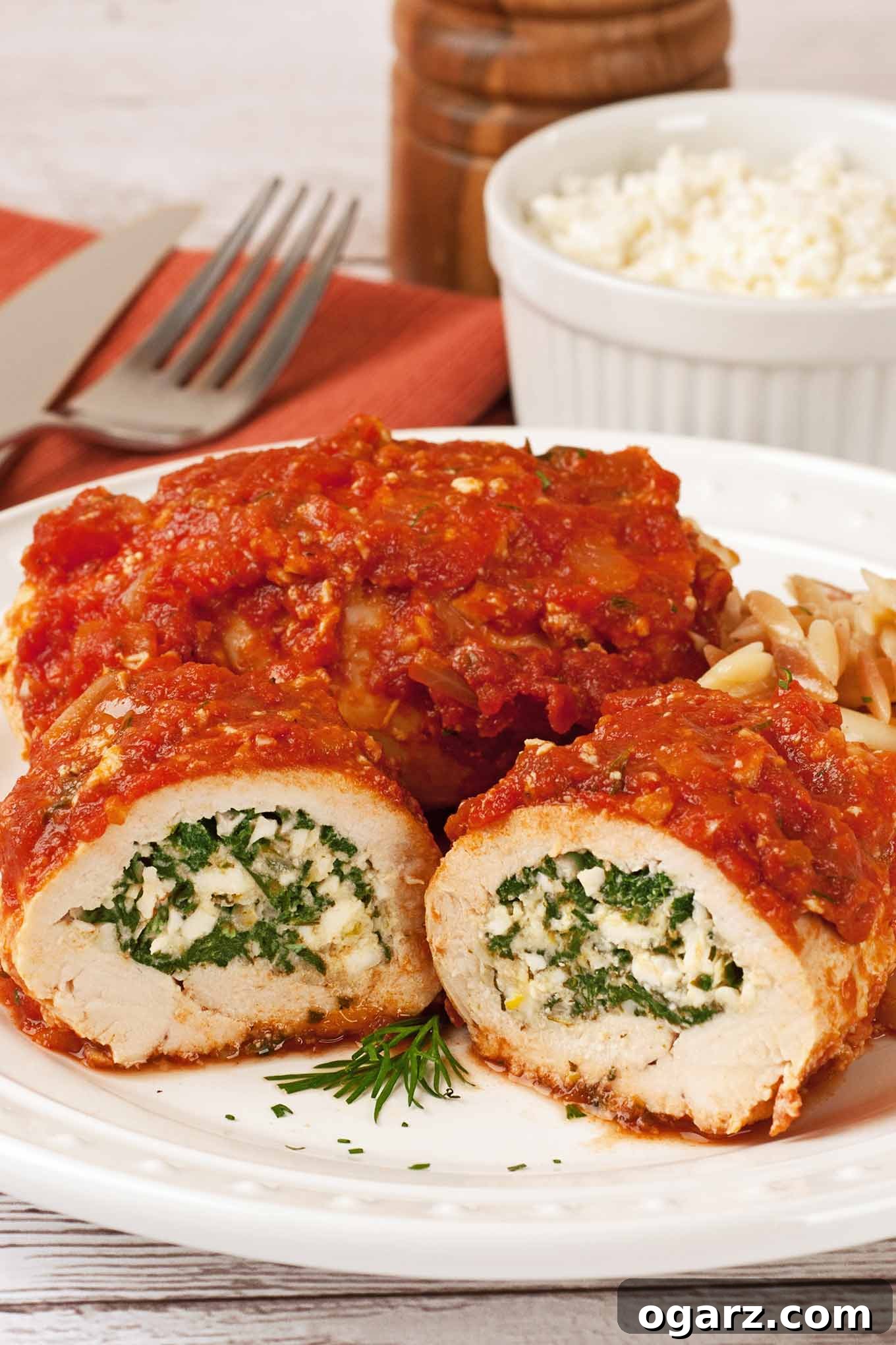 Greek-Inspired Feta and Spinach Stuffed Chicken 2 A beautifully arranged dinner plate featuring two golden-brown, boneless chicken breasts, expertly rolled and generously stuffed with a vibrant mixture of fresh spinach and savory feta cheese. The chicken is artfully drizzled with a rich, Greek-style tomato sauce. Accompanying the dish are gleaming utensils, a small bowl of crumbled feta cheese, and a rustic pepper mill in the soft-focused background, all hinting at a delicious, homemade meal.