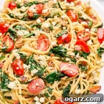 A serving platter of blue cheese pasta with caramelized onions, bacon, fresh spinach, and tomatoes, a block of blue cheese, fresh basil, and halved grape tomatoes in the background.