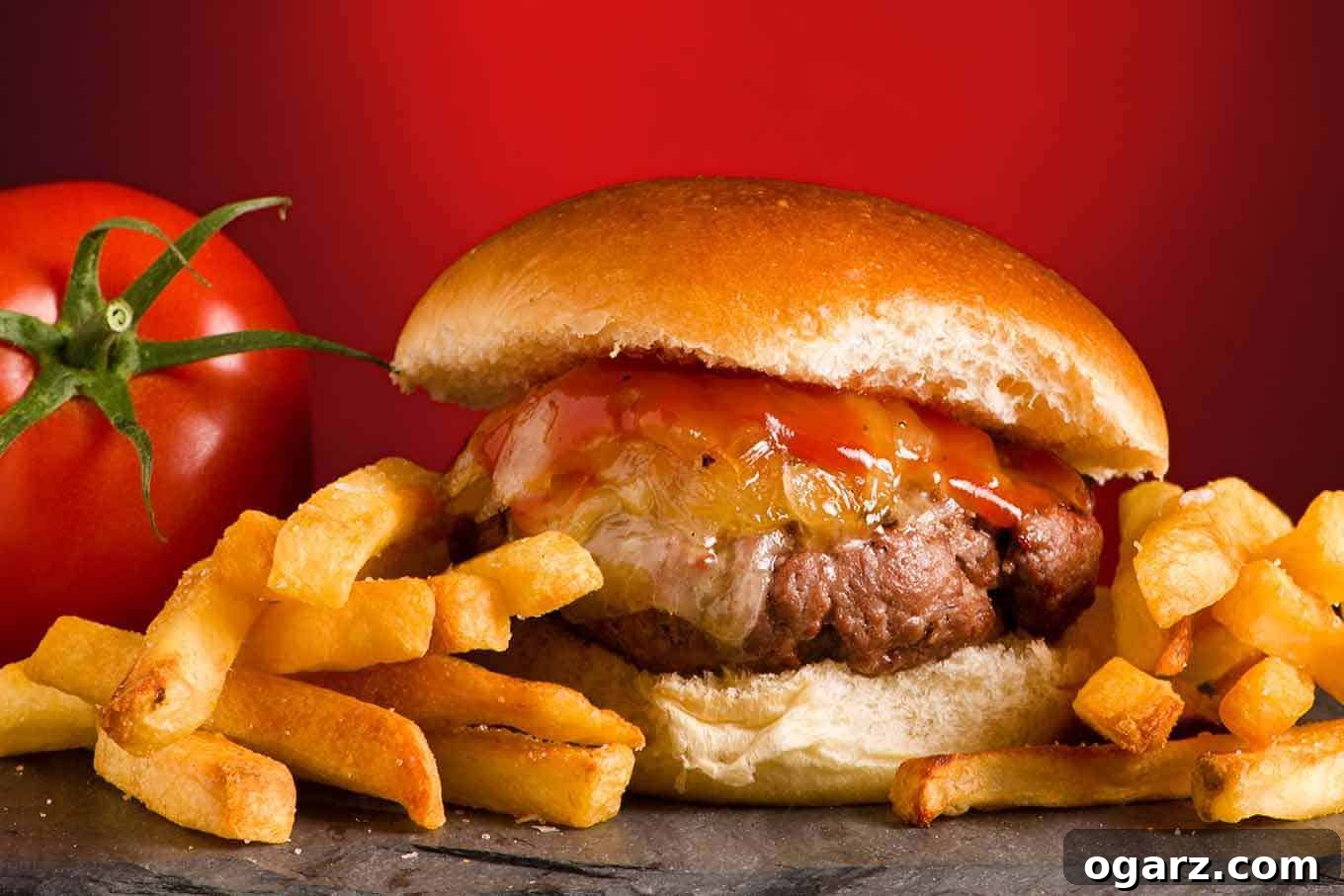 A close view of a grilled burger topped with cheddar cheese, maple ketchup and grilled onions on a slate board, fries to the side.