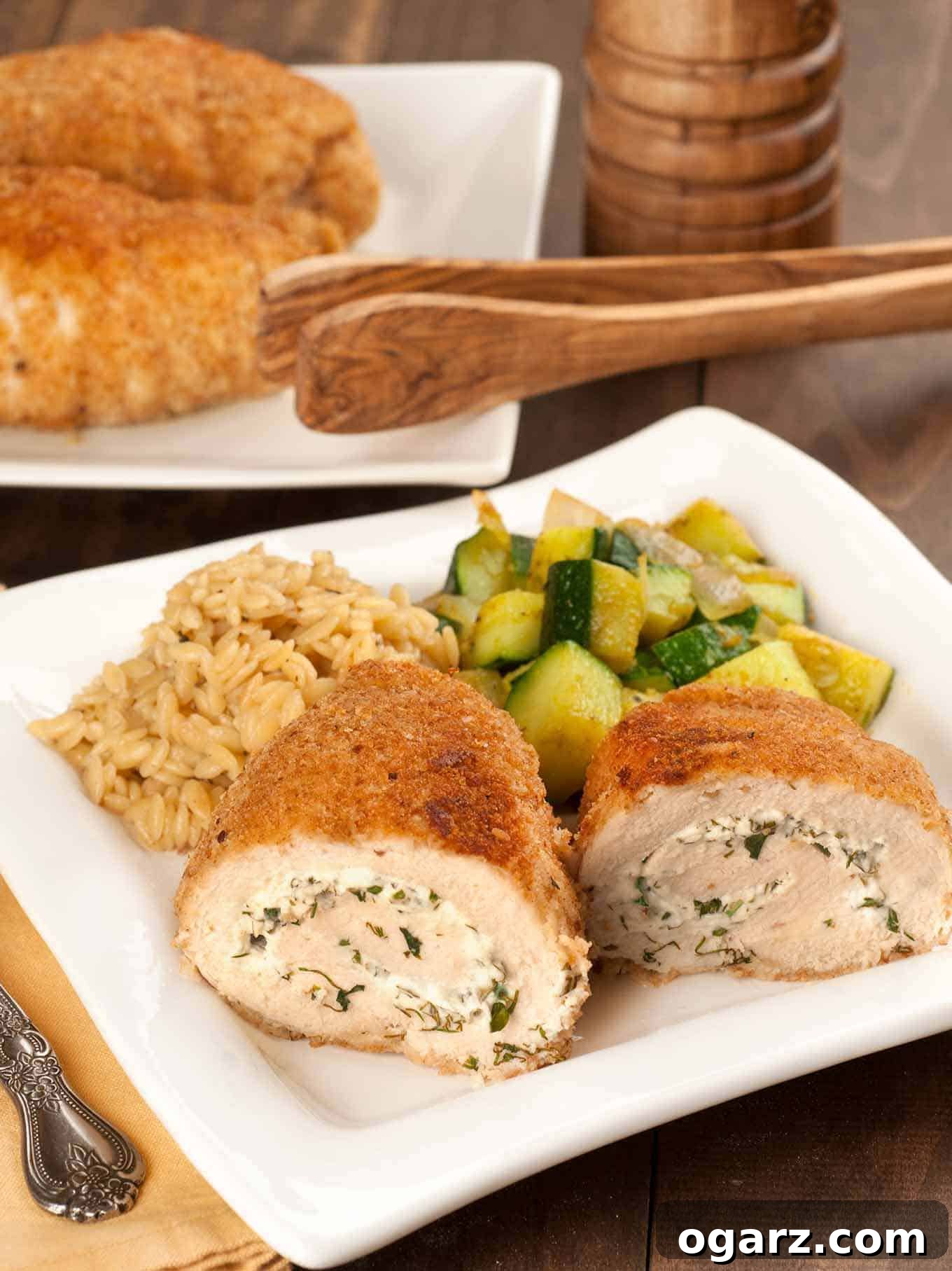 Herbed Goat Cheese-Filled Chicken Pockets 2 Rolled, crumb-coated chicken breasts filled with herbed goat cheese, served with sautéed zucchini and orzo on a square white plate, more chicken in the background.