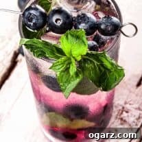 A highball glass filled with a blueberry mojito, muddled berries at the bottom, and a skewer of blueberries and mint sprig as garnish.