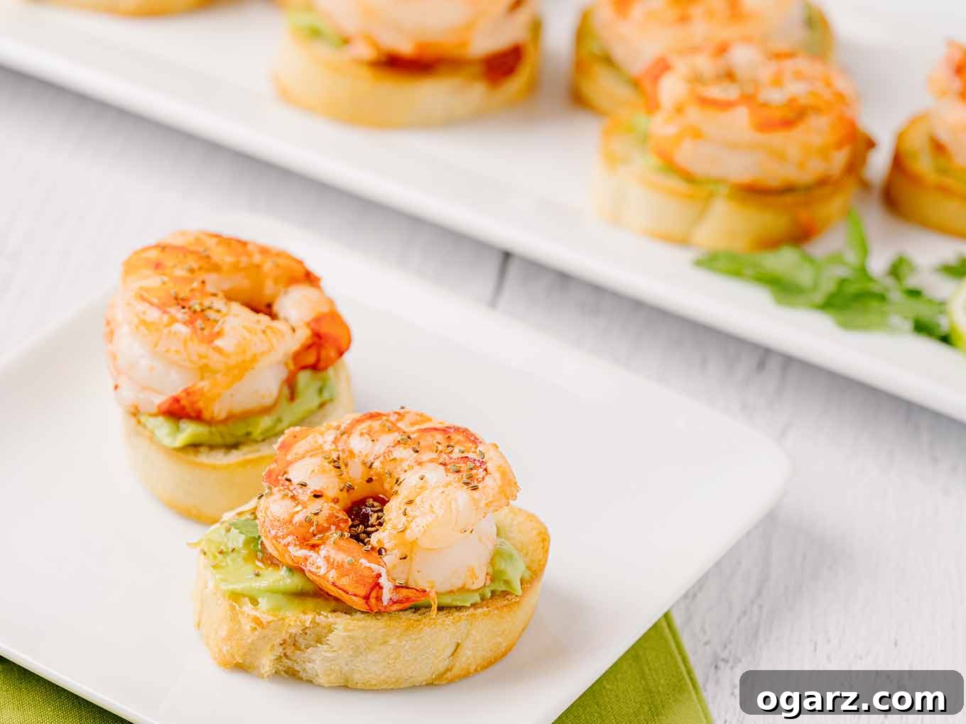 A close-up of two elegantly plated shrimp crostini on a small white appetizer plate, with the larger serving platter subtly blurred in the background, emphasizing individual portion presentation.