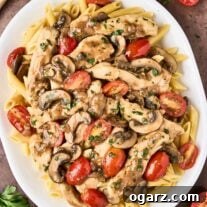 An overhead view of chicken Marsala pasta on a white oval serving platter, surrounded by a scattering of halved grape tomatoes, a sprig of parsley, and a wooden spoon.
