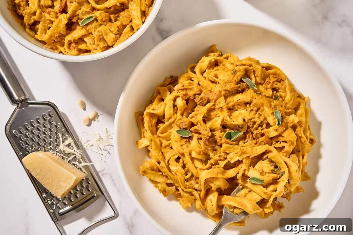 Homemade creamy pumpkin pasta in white bowls with crispy pancetta, sage, and toasted pine nuts, a cheese grater and chunk of Parmesan to the side.