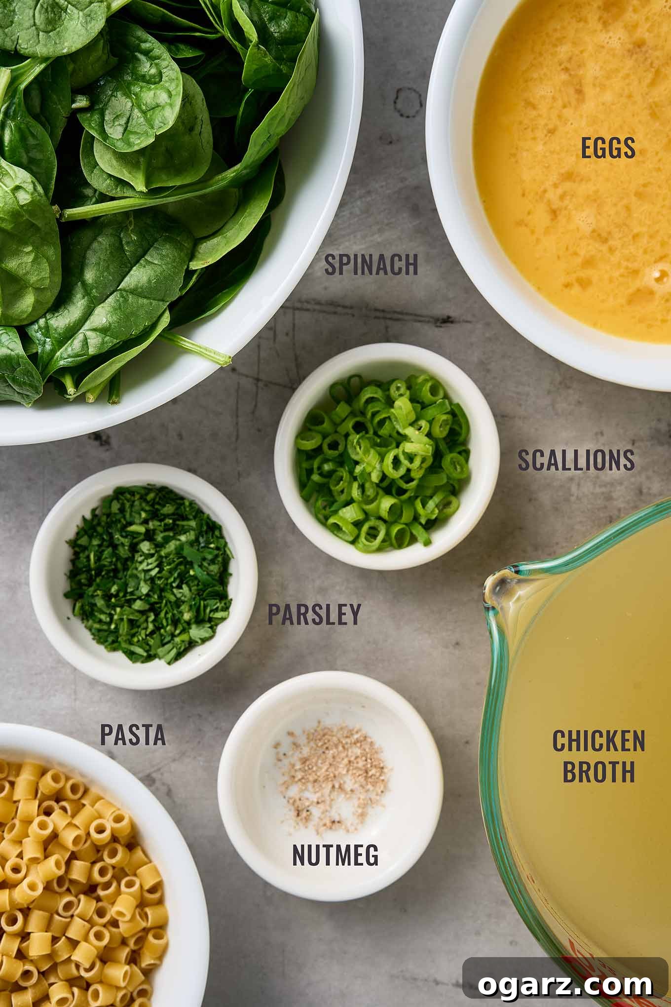 Spinach Pasta Stracciatella Soup 3 Labeled overhead view of ingredients for stracciatella soup, including spinach, eggs, scallions, parsley, pasta, nutmeg, and chicken broth.