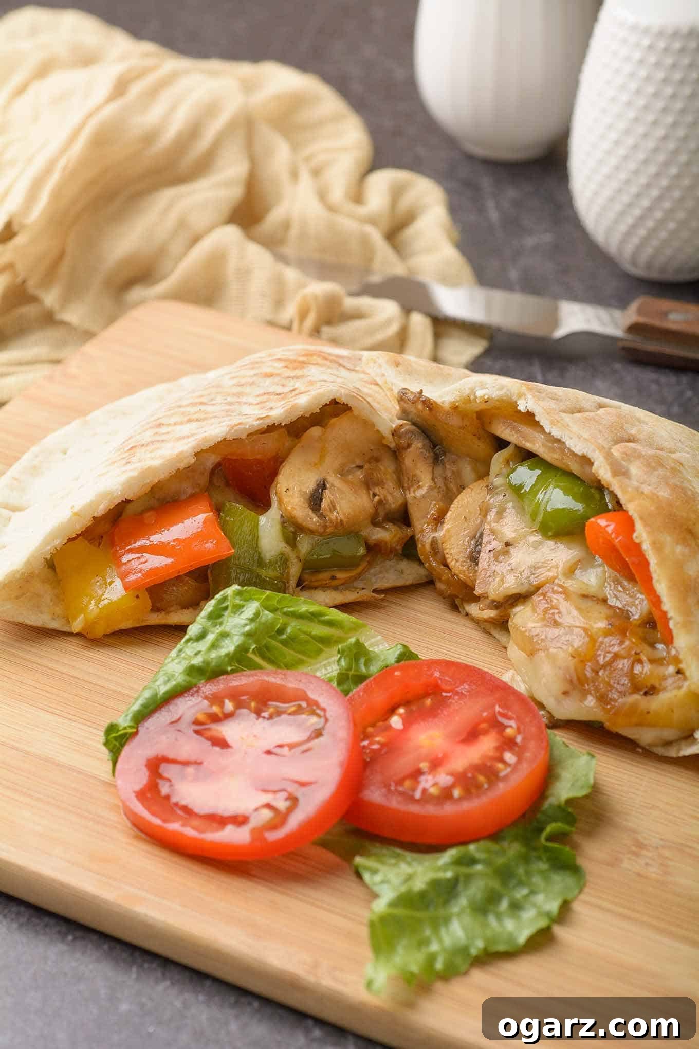 Pita bread stuffed with sautéed peppers and mushrooms, melted Muenster cheese, lettuce, and tomatoes on a cutting board, knife and cloth napkin in the background.