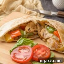 Pita bread stuffed with sautéed peppers and mushrooms, melted Muenster cheese, lettuce, and tomatoes on a cutting board, knife and cloth napkin in the background.