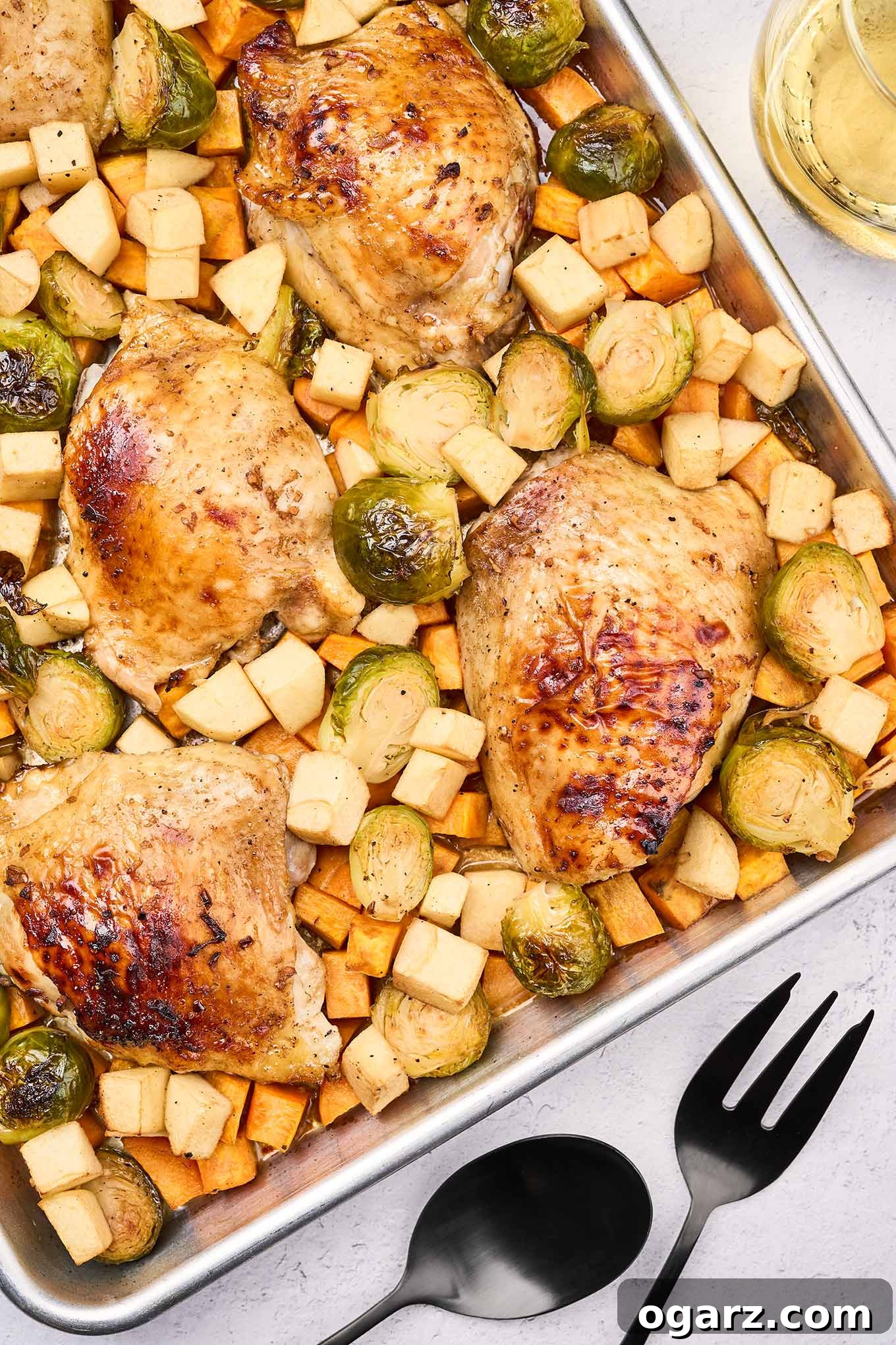 One-Pan Chicken and Seasonal Veggies 2 An overhead view of sheet pan chicken with seasonal vegetables next to black serving utensils and a glass of white wine. A honey-balsamic glazed chicken thigh is surrounded by roasted Brussels sprouts, sweet potatoes, and apples.