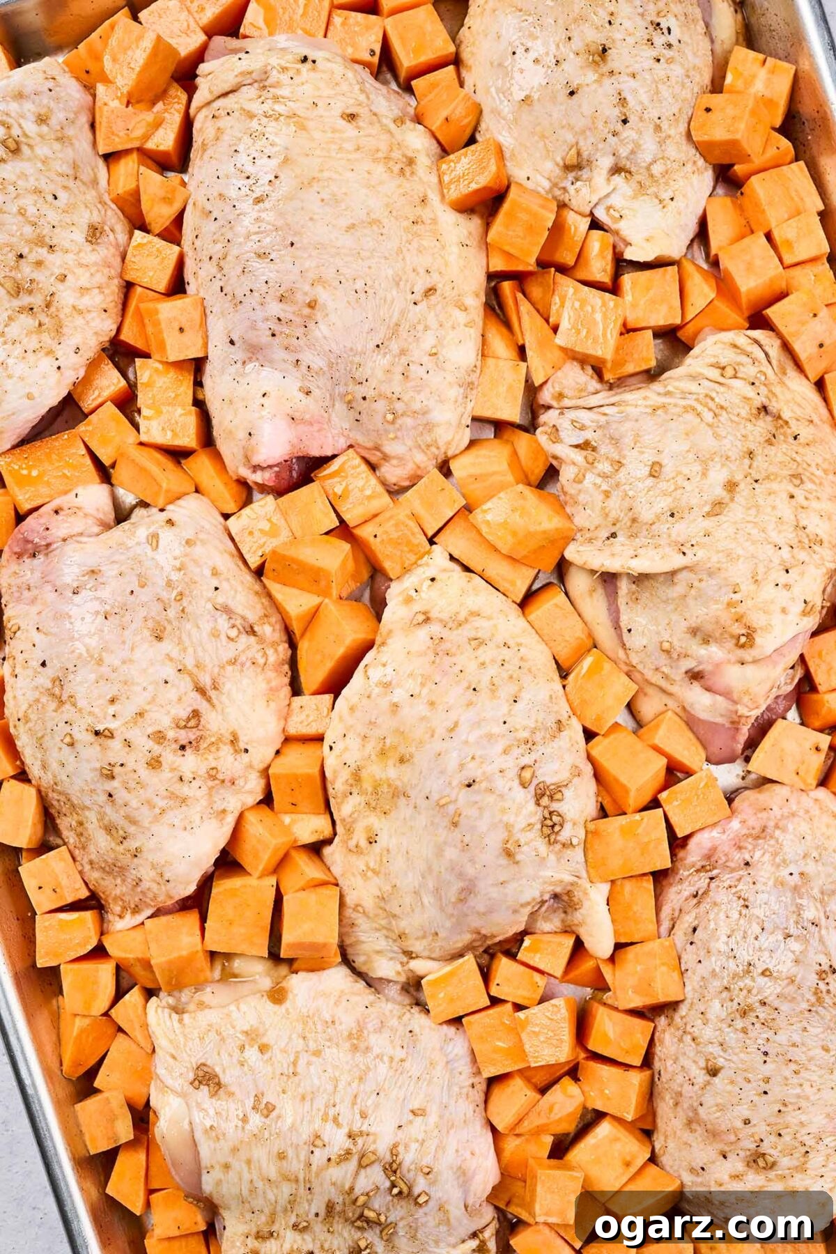 One-Pan Chicken and Seasonal Veggies 7 A sheet pan filled with raw chicken thighs, seasoned and surrounded by chunks of sweet potatoes, ready for oven-roasting. The ingredients are evenly spaced.