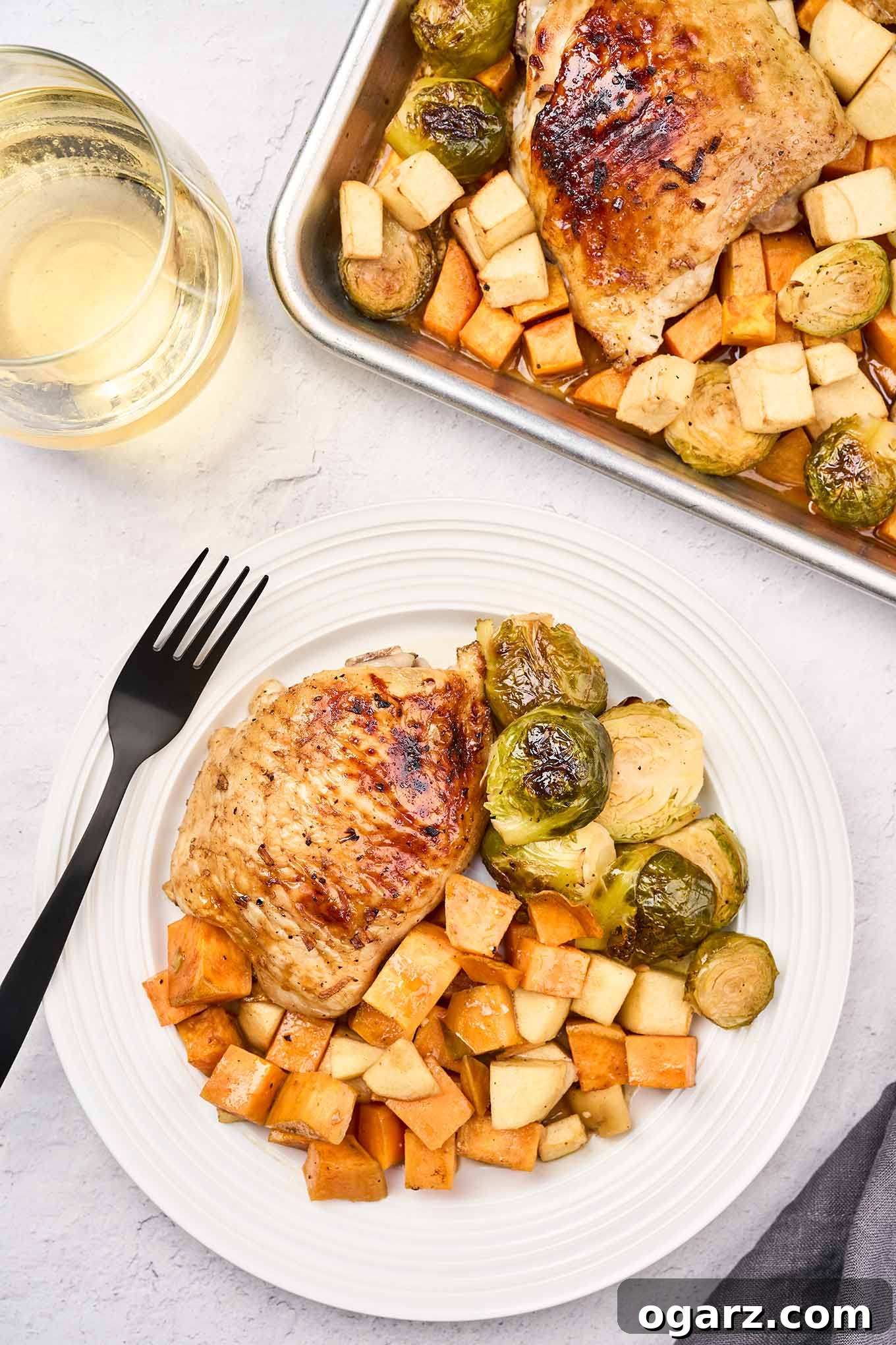 One-Pan Chicken and Seasonal Veggies 9 A plate with a honey-balsamic glazed roasted chicken thigh, sweet potatoes, apples, and Brussels sprouts, next to a fork, a glass of wine and the sheet pan with more. The meal looks inviting and delicious.