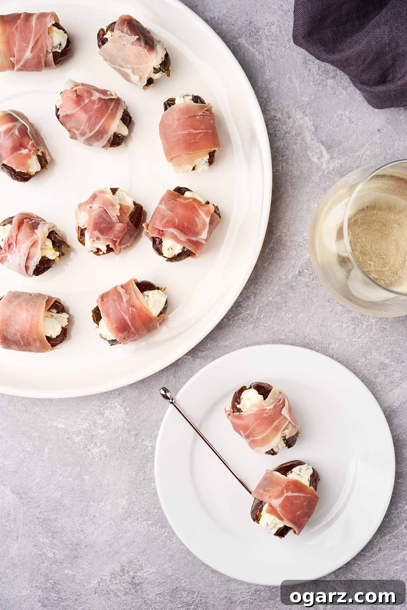 An overhead view of a serving plate of prosciutto-wrapped stuffed dates, a glass of white wine, and an appetizer plate with two stuffed dates and a silver cocktail pick.