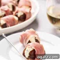 Stuffed dates wrapped in prosciutto on an appetizer plate with a silver pick, a glass of white wine and serving plate in the background.