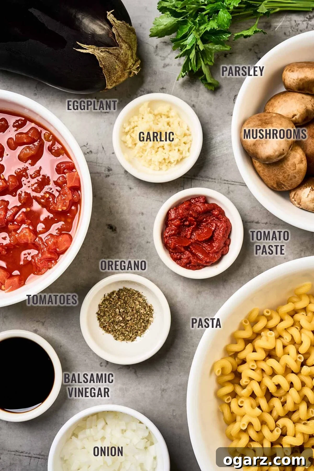 Umami Eggplant and Mushroom Pasta 3 A labeled, overhead view of the ingredients needed to make eggplant and mushroom pasta; eggplant, pasta, mushrooms, onion, garlic, parsley, tomato paste, oregano, balsamic vinegar, and tomatoes.