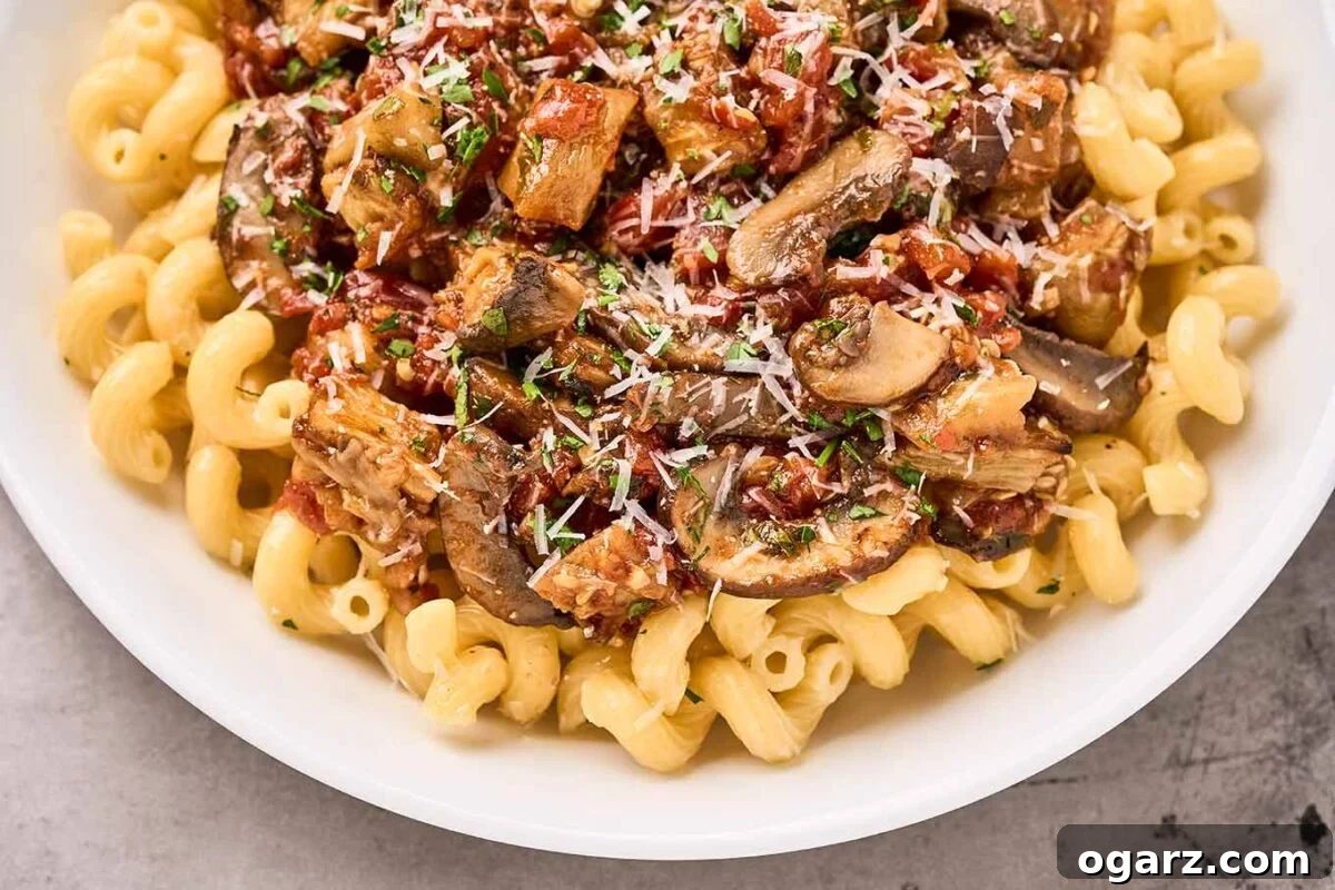 Umami Eggplant and Mushroom Pasta 6 Close overhead view of a bowl of cavatappi pasta topped with a roasted eggplant, mushroom, and tomato sauce, with grated Parmesan cheese and chopped parsley for garnish.