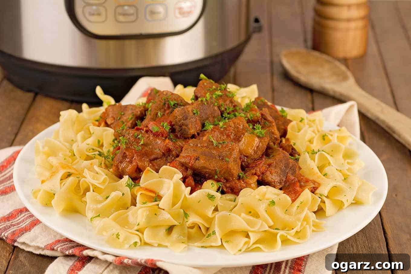 Hearty Hungarian Beef Goulash Pressure Cooked to Perfection 3 A close view of a plate of beef goulash served over egg noodles and garnished with chopped parsley, a wooden spoon, pepper mill, and Instant Pot in the background.