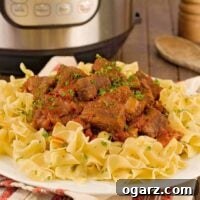 A close view of a plate of beef goulash served over egg noodles and garnished with chopped parsley, a wooden spoon, pepper mill, and Instant Pot in the background.
