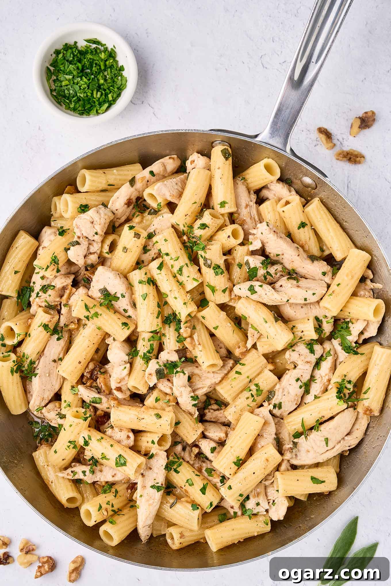 Nutty Brown Butter Chicken Pasta 9 An overhead view of a skillet filled with brown butter pasta and tender strips of chicken, garnished with fresh parsley. A small prep bowl with more chopped parsley sits to the side, ready for serving.