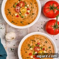 Two bowls of vibrant gazpacho soup garnished with fresh vegetables and parsley, surrounded by ingredients like tomatoes, cucumbers, and bell peppers.