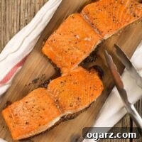 Two grilled salmon fillets on a cedar plank on top of a kitchen towel, metal tongs to the side.