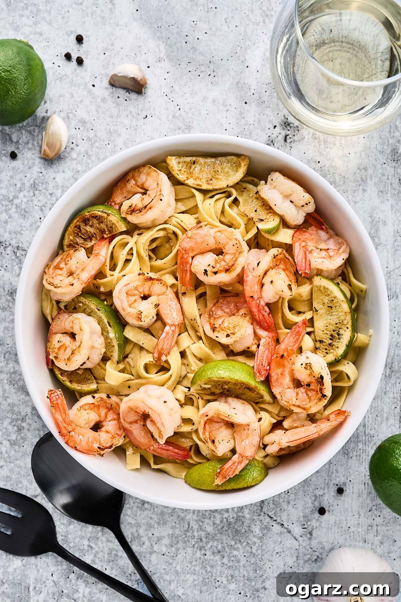 Zesty Garlic Lime Shrimp Pasta 6 Garlic lime shrimp pasta garnished with lime wedges in a white bowl on a light stone surface, surrounded by garlic cloves, peppercorns, limes, and white wine.