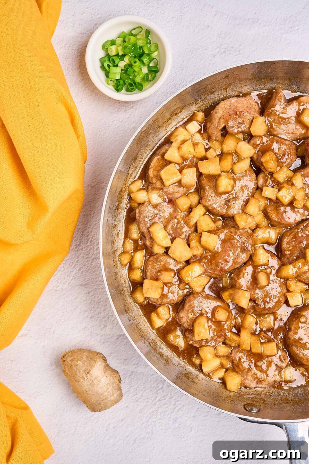 Tropical Pineapple Pork Skillet 4 A skillet filled with tender pork medallions, and pineapple chunks in a sweet-savory rum sauce, a small bowl of chopped scallions and a knob of ginger to the side, illustrating the cooking process.
