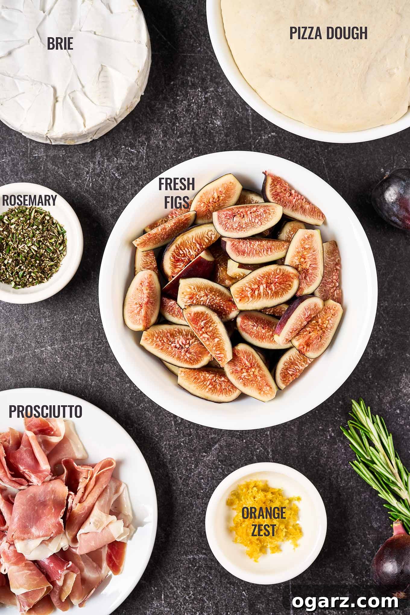 A beautifully arranged overhead view of all the essential ingredients for making fig and prosciutto pizza. This includes fresh, ripe figs, a ball of ready-to-use pizza dough, thinly sliced prosciutto, fragrant rosemary sprigs, bright orange zest, and creamy Brie cheese.