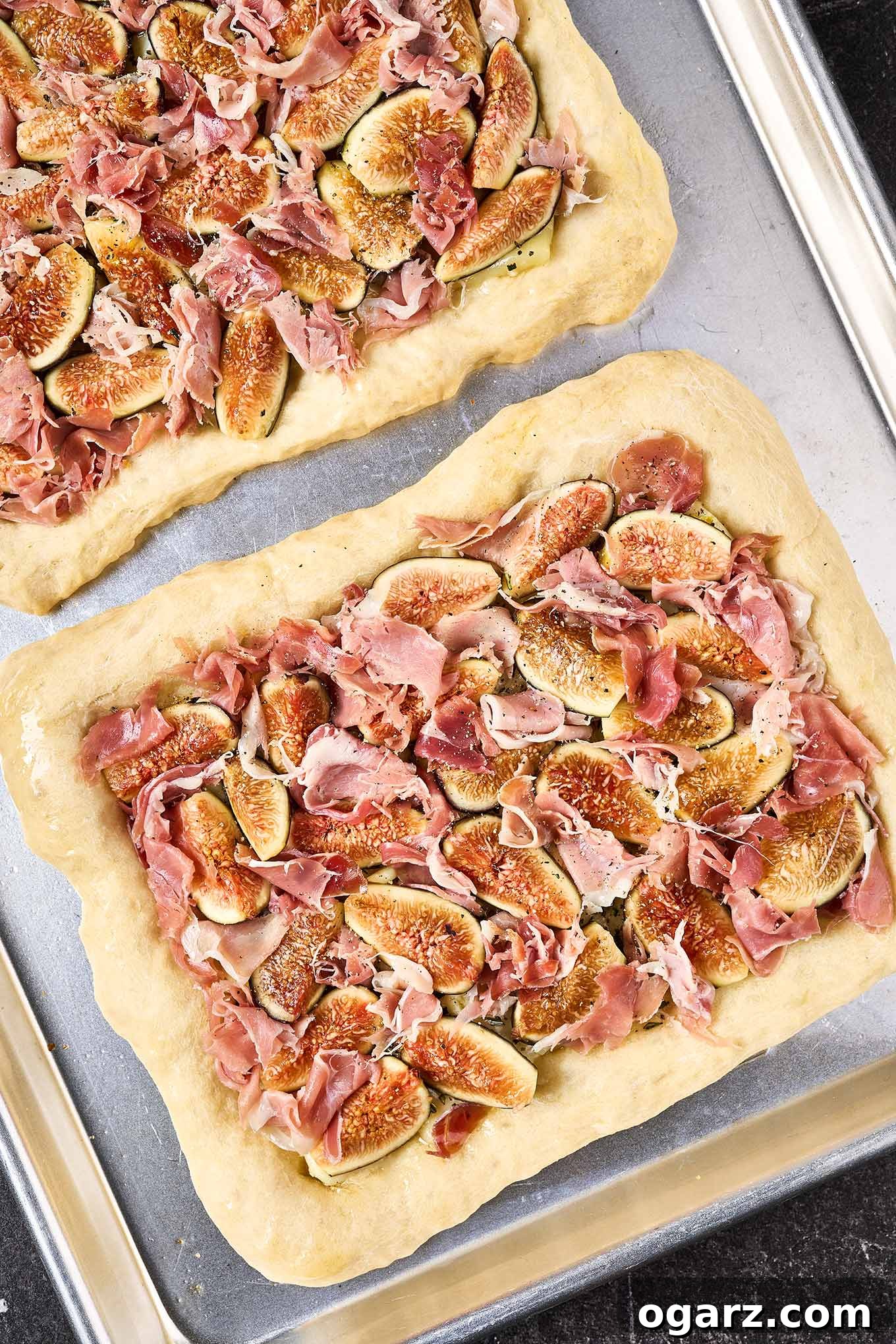 A detailed recipe preparation photo showcasing two pre-baked pizza crusts, artfully topped with generous dollops of creamy Brie, vibrant quartered figs, and savory pieces of prosciutto, perfectly arranged and ready for their final bake in the oven.