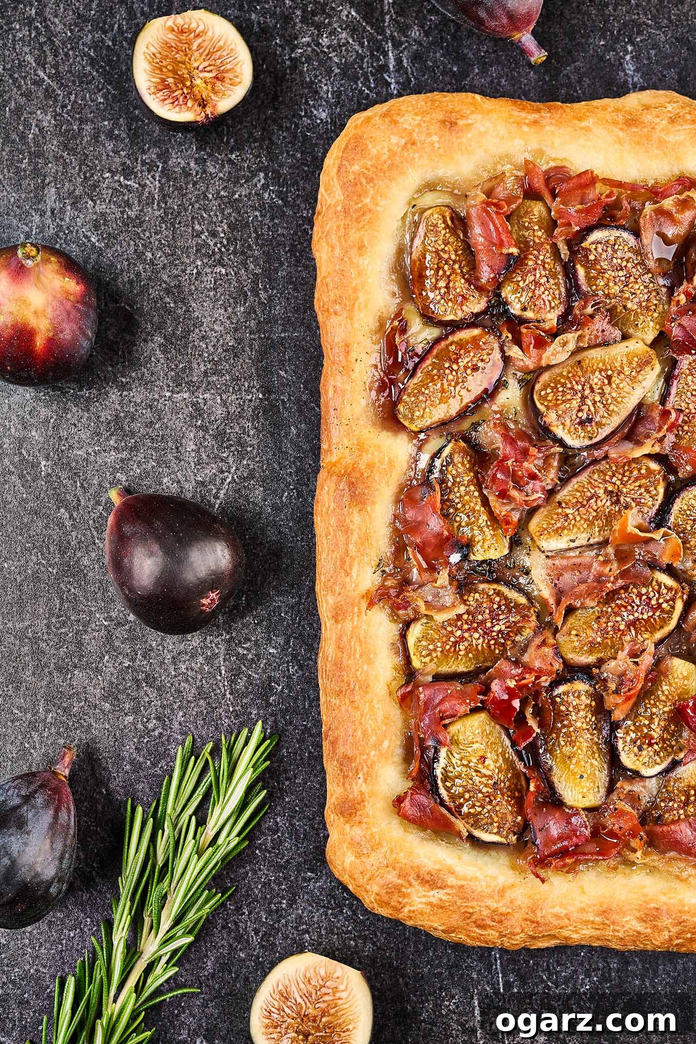 A freshly baked fig and prosciutto pizza, golden brown and bubbling, resting on a rustic slate background. It is accompanied by fresh, ripe whole figs and delicate sprigs of aromatic rosemary, highlighting its gourmet ingredients and inviting presentation.