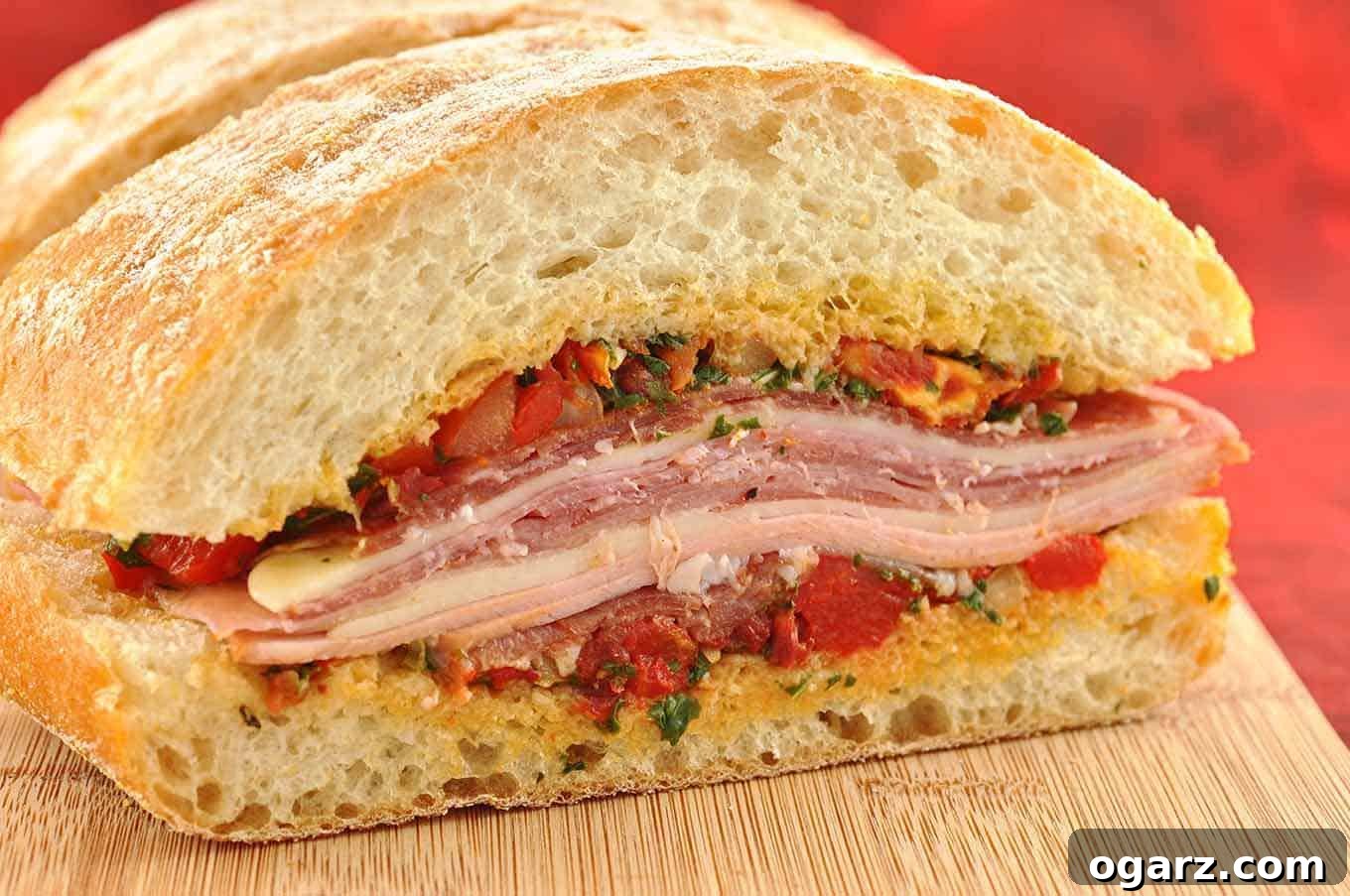 Zesty Mediterranean Muffuletta 2 Close-up of a Mediterranean Muffuletta sandwich, beautifully layered with cured Italian meats, Asiago cheese, and a vibrant basil, tomato, and roasted pepper relish, all nestled within a crusty ciabatta bread.