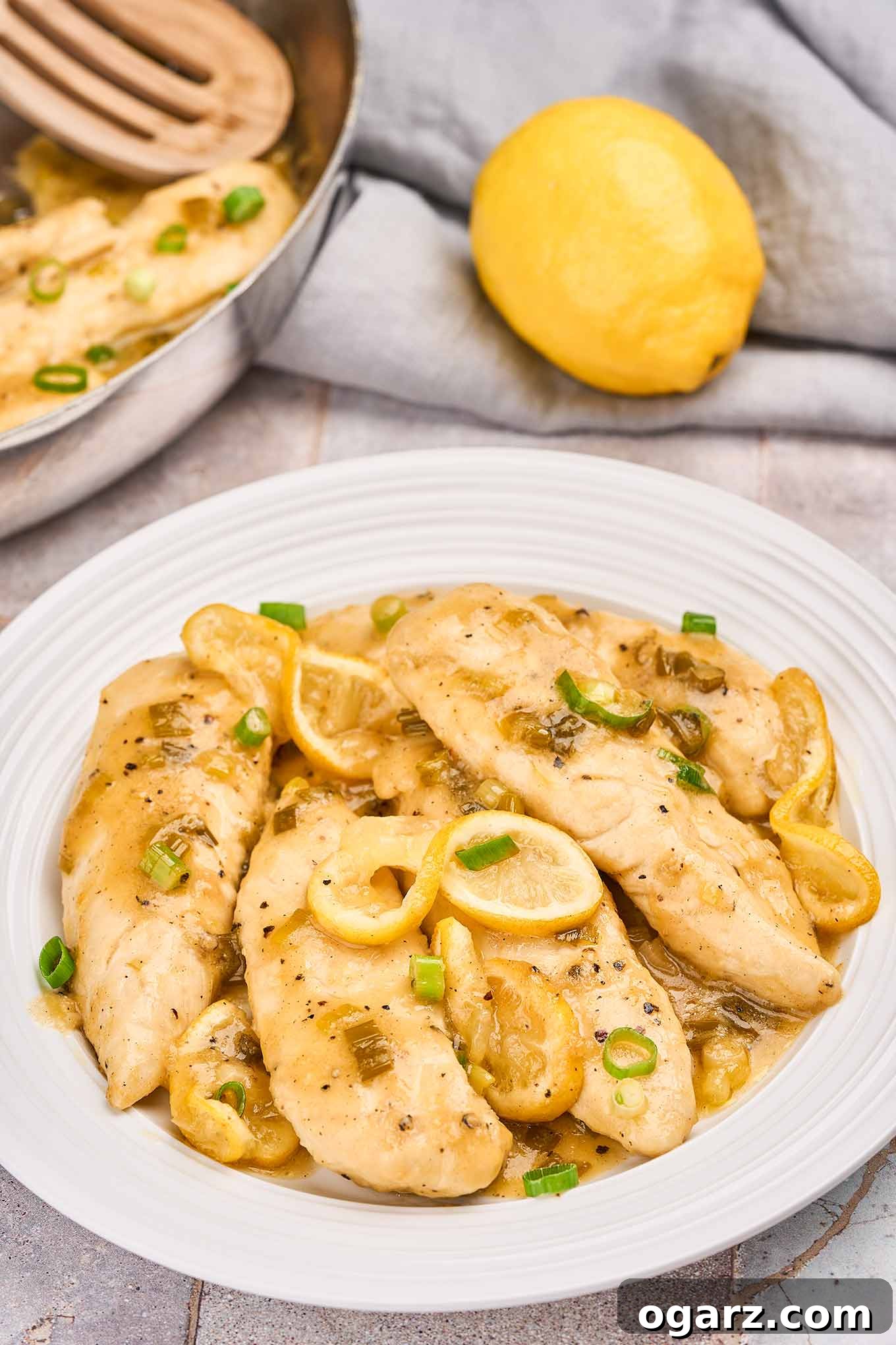 A shallow white serving bowl filled with vibrant lemon pepper chicken tenders in a glossy pan sauce, garnished with fresh lemon slices and chopped green scallions, suggesting a quick and delicious weeknight meal.