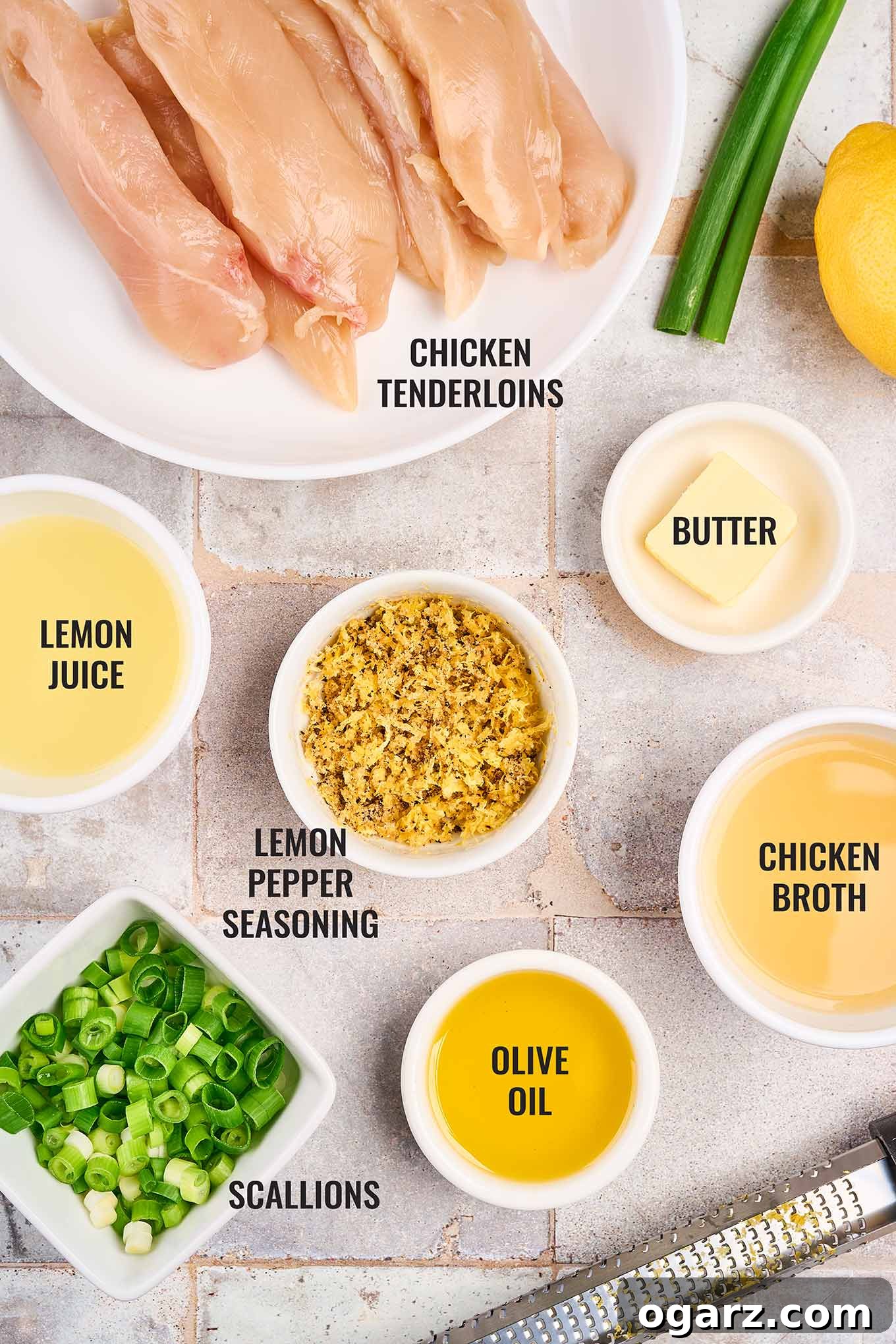 A collection of fresh ingredients laid out on a tiled kitchen counter, including raw chicken tenderloins, fresh lemons, a stick of butter, a bottle of olive oil, a bowl of flour, chicken broth, and a small dish of homemade lemon pepper seasoning blend.