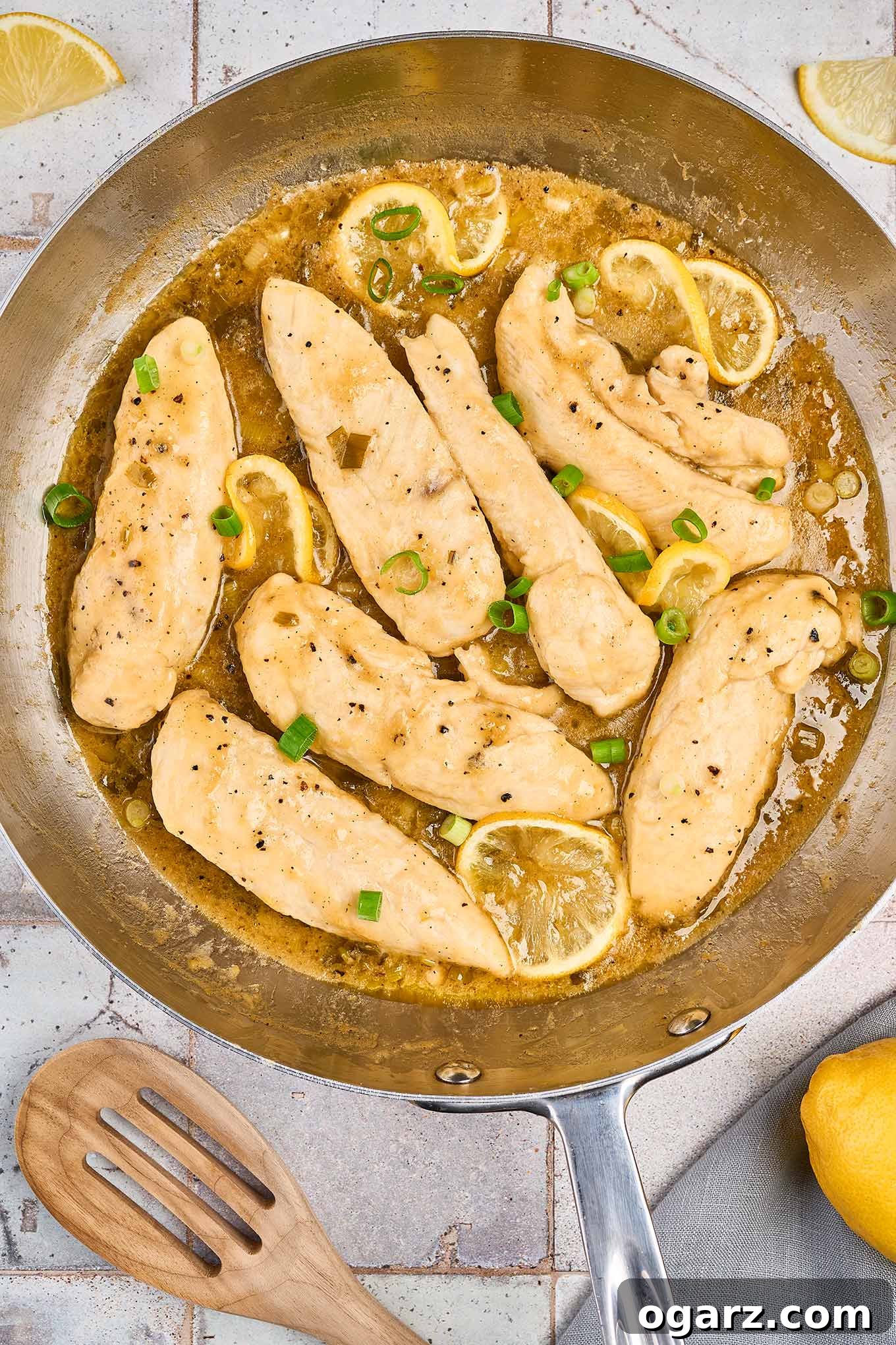 Close-up shot of pan-seared lemon pepper chicken tenderloins sizzling in a stainless steel frying pan, bathed in a glossy lemon pan sauce and garnished with fresh lemon slices and vibrant chopped scallions, ready to be served.