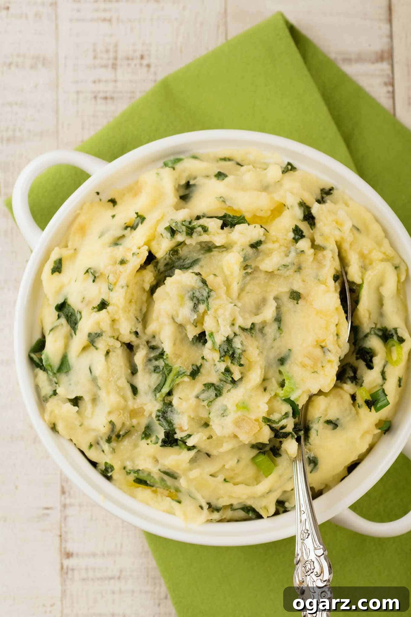 Ireland's Creamy Colcannon 2 An overhead view of a white casserole filled with creamy mashed potatoes mixed with sautéed kale, sweet onion, and scallions (colcannon), and a serving spoon.