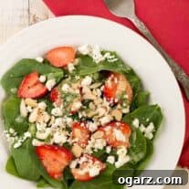 An overhead view of a composed salad of baby spinach, sliced fresh strawberries, crumbled goat cheese, toasted slivered almonds, topped with basil vinaigrette.