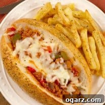 A plate with a sub roll filled with a sloppy-Joe style mix of crumbled Italian sausage, green pepper, tomato sauce, and melted mozzarella, fries on the side.