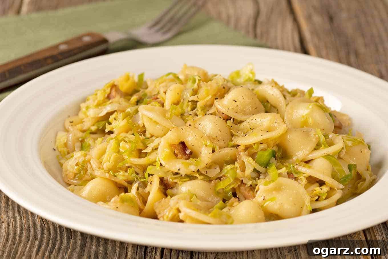 A close-up shot of a white plate filled with perfectly portioned orecchiette pasta, generously tossed with golden, shredded Brussels sprouts and crispy bacon bits, ready to be enjoyed. A fork and a folded white napkin are visible in the background.