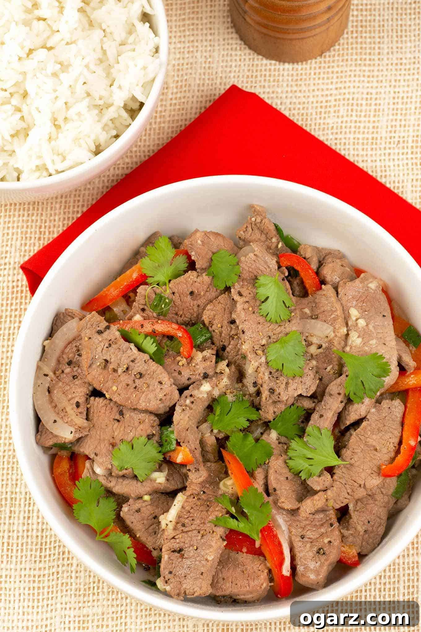 Bold Vietnamese Garlic & Black Pepper Stir-Fry 2 A vibrant bowl of Vietnamese-style garlic and black pepper stir-fry, featuring tender slices of beef, lamb, or pork, vibrant red bell pepper strips, and sweet onion, garnished generously with fresh cilantro leaves, accompanied by a side of fluffy white rice.