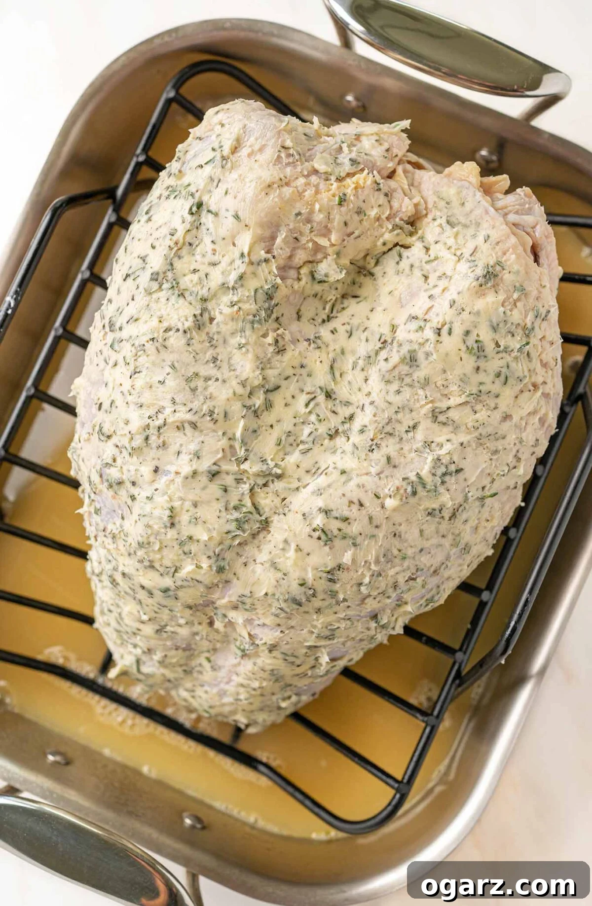A turkey breast on a rack inside a roasting pan, coated in herb butter and ready to begin roasting.