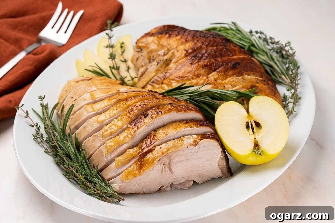 Carved turkey breast on a platter garnished with fresh rosemary and thyme and half an apple, serving fork in the background, ready for a delicious meal.