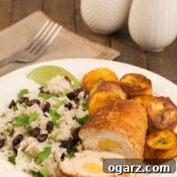 A dinner plate with a crumb-coated, rolled boneless chicken breast stuffed with sweet plantains, served with black beans and rice and sautéed maduros, a fork, napkin, and salt and pepper shakers in the background. Close-up of golden brown plantain stuffed chicken.