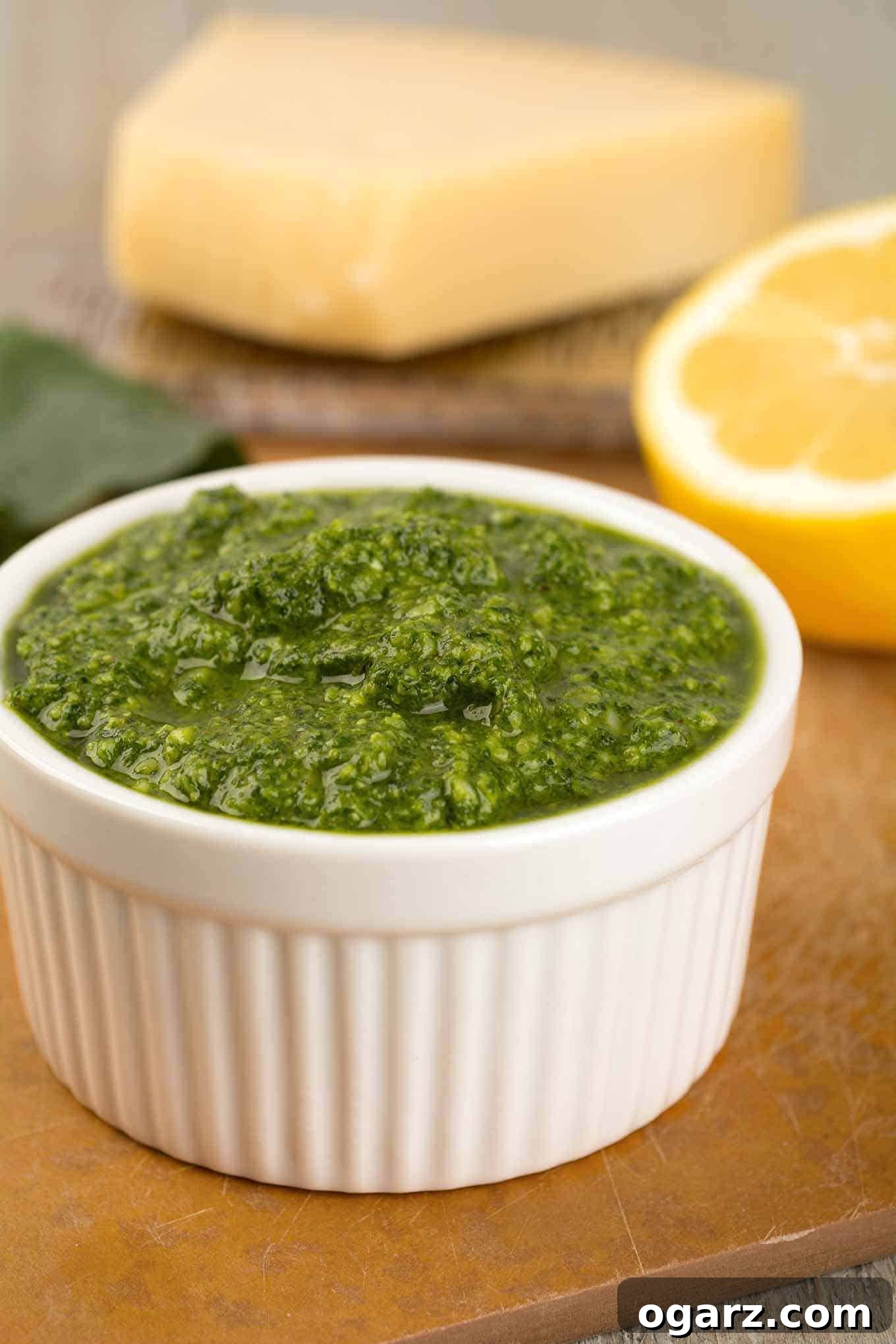 A small white bowl filled with vibrant green kale pesto, accompanied by a halved lemon and a rustic cutting board featuring a wedge of Parmesan cheese.