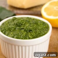 A small white bowl filled with kale pesto with a half lemon and cutting board with a wedge of Parmesan cheese in the background.