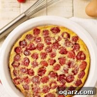 An overhead view of freshly baked raspberry clafoutis dusted with powdered sugar in a white pie plate, surrounded by a whisk, eggs, and fresh raspberries.