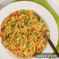 An overhead view of a bowl of stir fried riced cauliflower combined with red bell pepper, scallions, scrambled egg, ginger, garlic, and cilantro, a wooden-handled fork to the right.