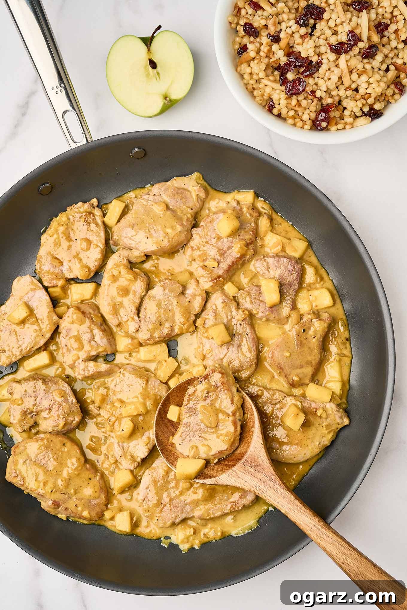 A skillet of pan seared pork tenderloin medallions and chunks of sautéed apple in a mild, creamy curry sauce, being stirred with a wooden spoon. A bowl of pearl couscous for serving in the background.