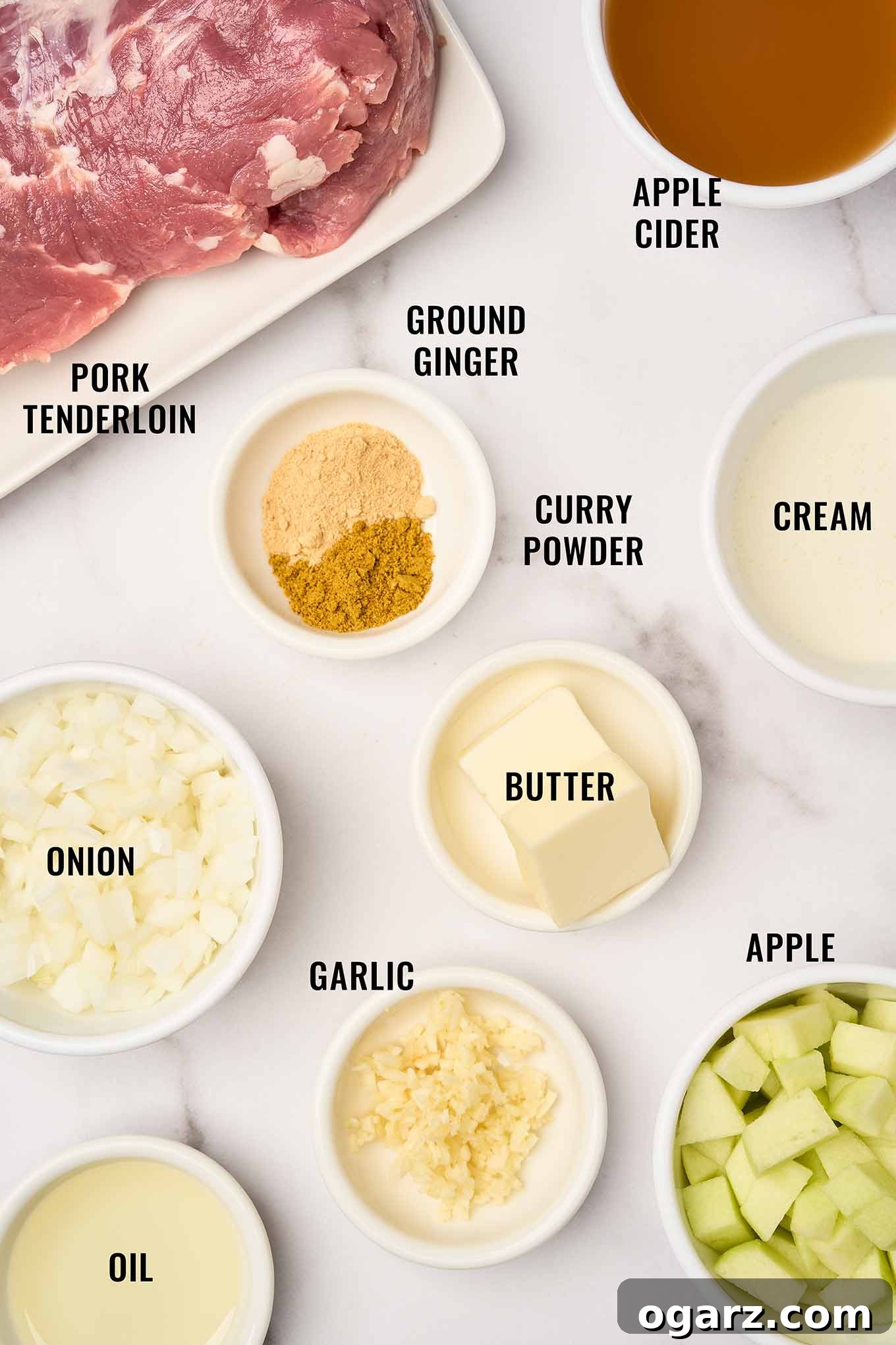 A labeled, overhead view of the ingredients needed to make pork curry with apples; pork tenderloin, apple cider, curry powder, ground ginger, onion, garlic, butter, cream, apple, and oil.