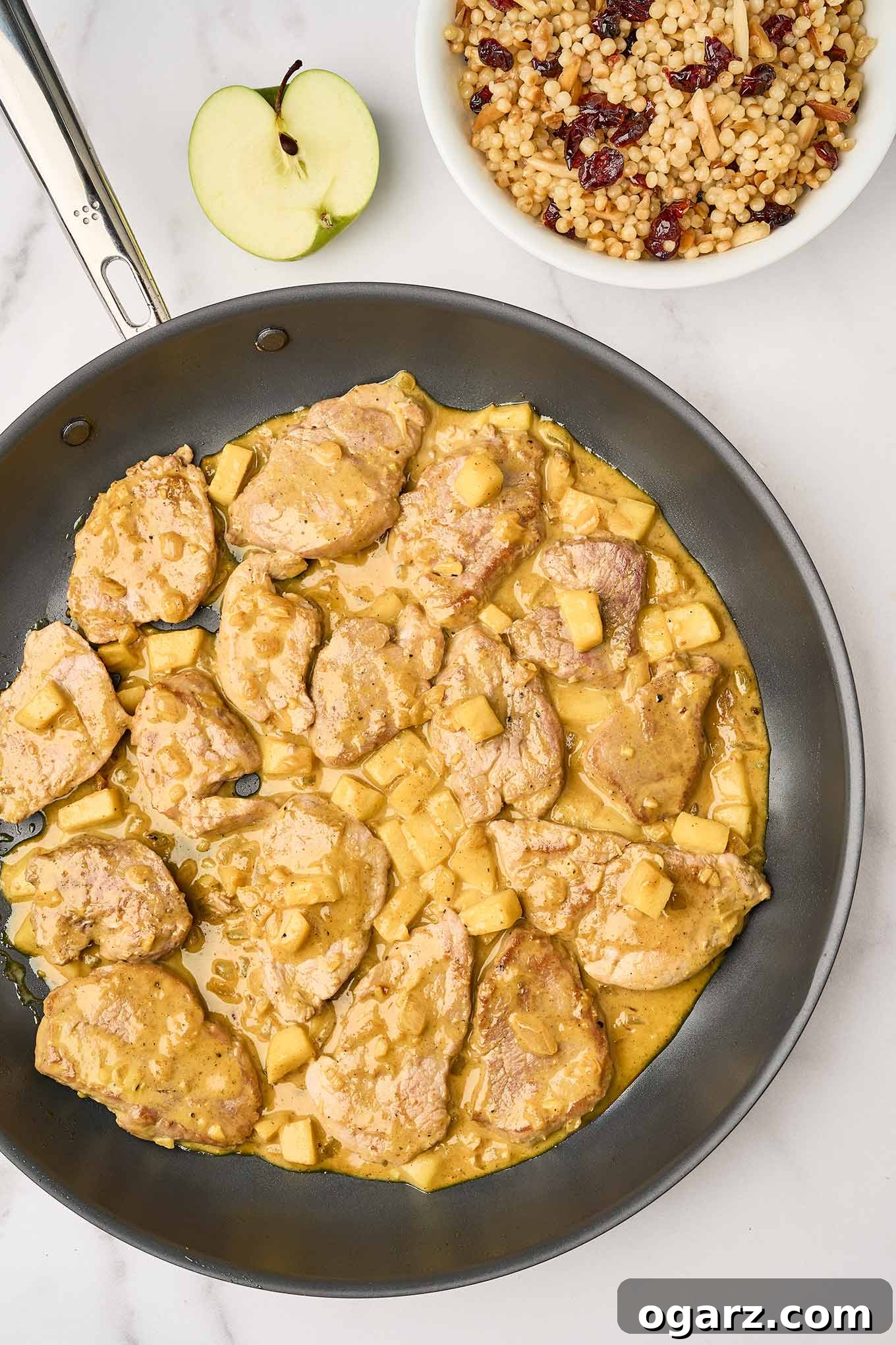 A skillet of pork curry with apples, coated in a light, creamy curry sauce, a bowl of toasted couscous for serving in the background, alongside a tart green apple cut in half.