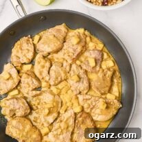 A skillet of pork curry with apples, coated in a light, creamy curry sauce, a bowl of toasted couscous for serving in the background, alongside a tart green apple cut in half.