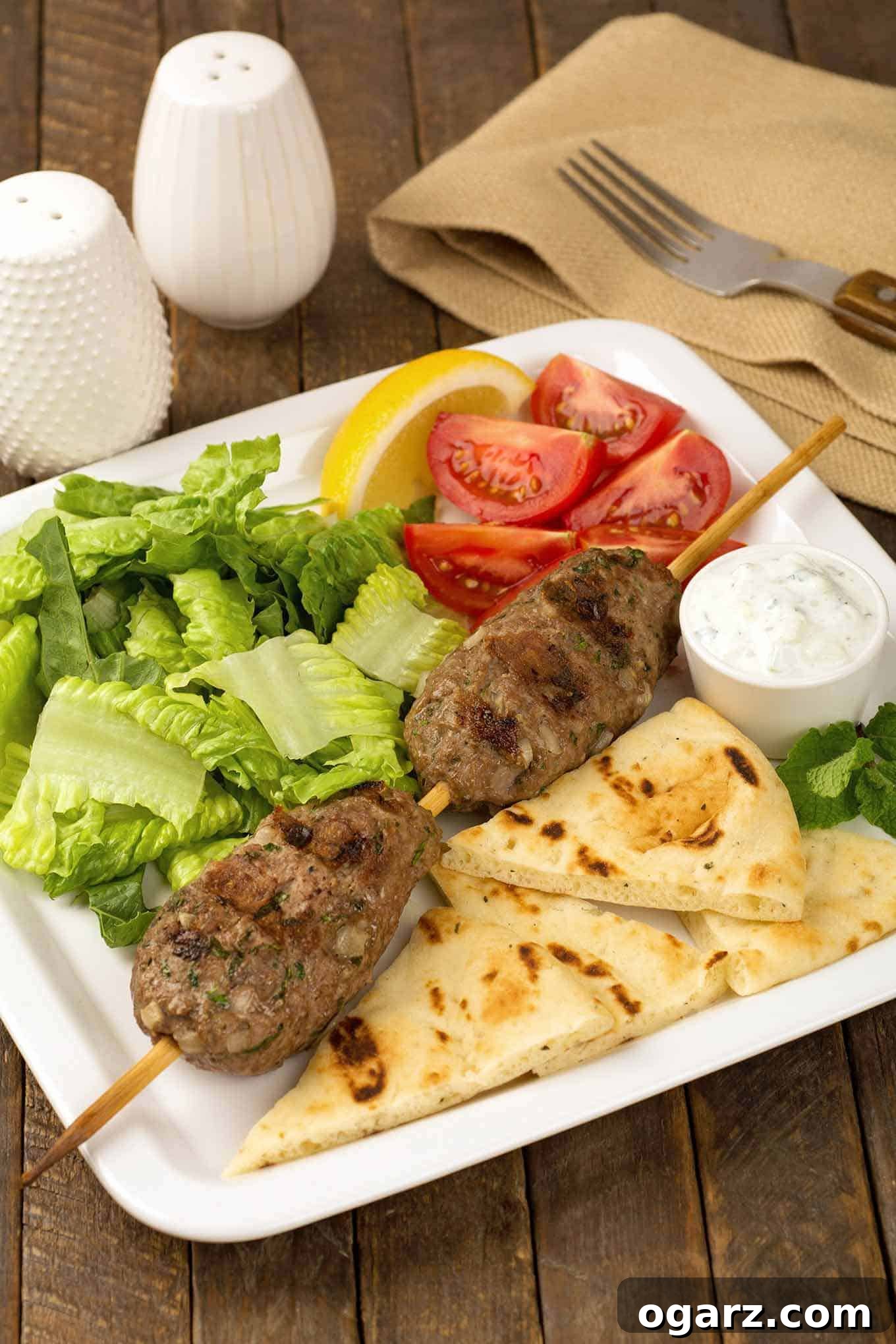 Flame-Grilled Moroccan Lamb Skewers 2 A dinner plate with two grilled skewered lamb meatballs (kofta), grilled pita bread, a small bowl of tzatziki sauce, lemon wedges, lettuce, and tomatoes; a fork, napkin, and salt and pepper shakers in the background.