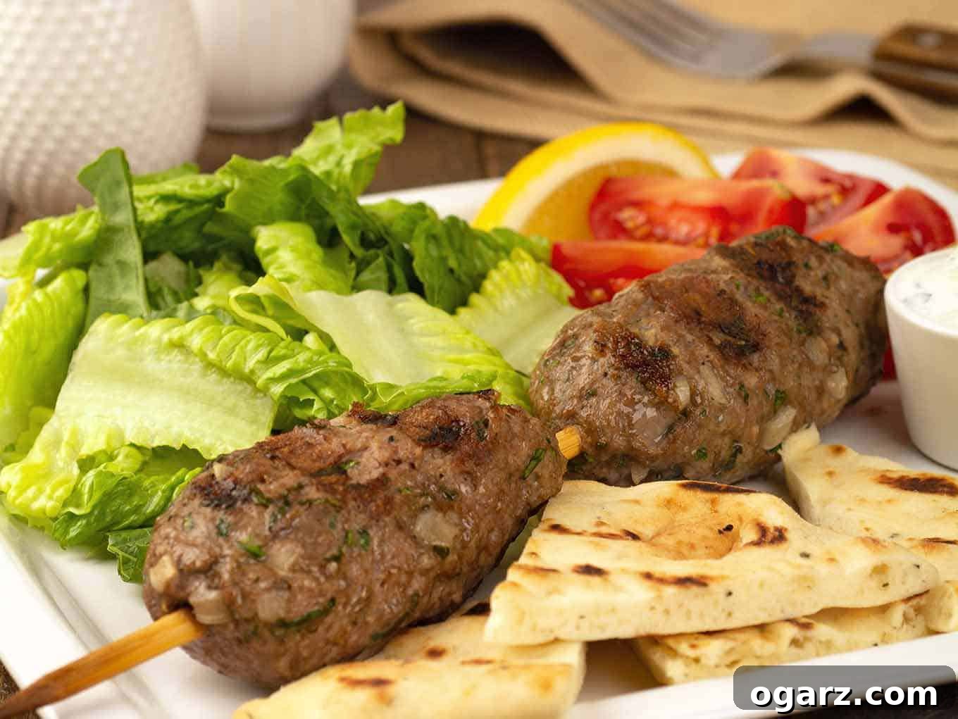 Flame-Grilled Moroccan Lamb Skewers 3 A close view of two grilled, skewered lamb meatballs, pita bread, lettuce, tomatoes, and lemon wedges on a dinner plate.